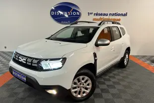 Vue 3/4 avant droite d'un Dacia Duster blanc 2023 avec feux allumés et barres de toit noires en intérieur showroom.