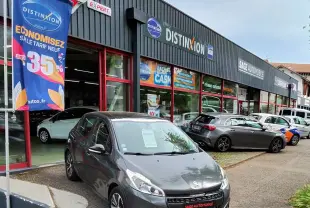 Peugeot 208 gris foncé métal vue 3/4 avant droit, garée devant un garage avec enseigne Sage Automobile.
