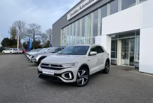 Volkswagen T-Roc gris en 3/4 avant droit, avec jantes alliage noires et calandre R-Line visible devant un concessionnaire.