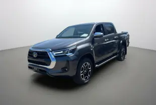 Toyota Hilux 4x4 gris atlas en 3/4 avant droit, avec jantes alu 18 pouces et marchepieds chromés visibles.