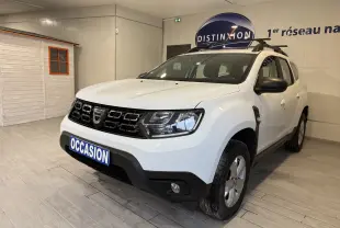 Dacia Duster blanc vue 3/4 avant droit en intérieur, avec calandre noire et barres de toit noires.