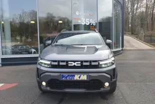 Vue avant d'un Dacia Duster 1.6 Hybrid gris schiste avec calandre noire et feux LED allumés, stationné devant un bâtiment vitré.