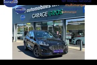 Ford Kuga 2024 noir Agate métallisé en 3/4 avant, avec calandre hexagonale et jantes alu sombres, devant un garage multimarque.