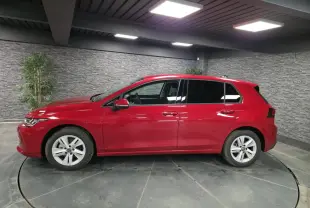 Profil droit d'une Volkswagen Golf VIII rouge métallisée, avec vitres arrière surteintées dans un showroom.
