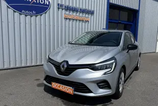 Renault Clio V gris en 3/4 avant droit, avec calandre noire et feux LED, stationnée devant un bâtiment métallique.