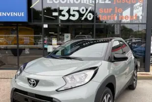 Toyota Aygo X 2025 gris clair en 3/4 avant droit, avec jantes alliage 17 pouces et feux LED, devant un showroom multimarque.