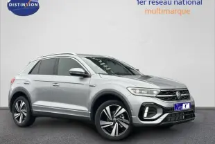Volkswagen T-Roc gris en 3/4 avant droit avec jantes alu et barres de toit visibles sur fond clair.