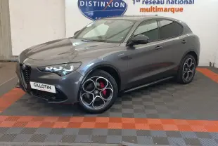 Alfa Romeo Stelvio gris foncé en 3/4 avant droit, avec jantes distinctives et phares LED allumés.