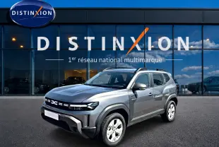 Dacia Duster 1.2 hybrid-g 150 gris schiste en 3/4 avant droit, avec barres de toit et jantes alu visibles.