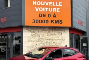 Renault Clio Evolution rouge vue en 3/4 avant droit, avec jantes noires et calandre distinctive, devant un bâtiment commercial.