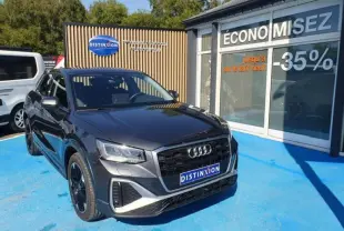 Audi Q2 30 TDI 116ch Design gris Manhattan métallisé vue 3/4 avant droit sur parking bleu devant vitrine promotionnelle.