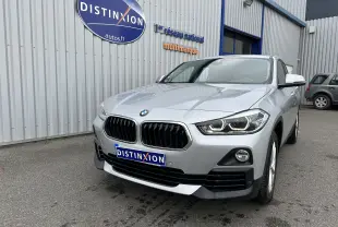 BMW X2 gris vue 3/4 avant avec calandre noire et phares LED allumés devant un bâtiment industriel