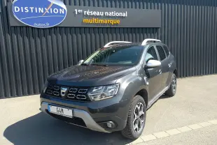 Dacia Duster gris Highland en 3/4 avant droit, avec jantes alliage et barres de toit blanches visibles.
