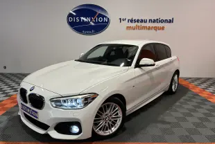 BMW Série 1 blanche vue en 3/4 avant droit, phares et feux de jour LED allumés dans un showroom.