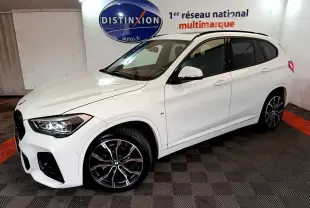 BMW X1 sDrive18i M Sport blanc vue 3/4 avant droit, jantes alliage noires et phares LED allumés, en intérieur showroom.