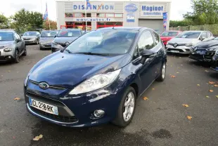 Ford Fiesta bleu foncé vue en 3/4 avant droit sur un parking de concession avec plusieurs autres véhicules autour.