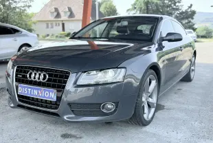 Audi A5 Sportback gris dauphin vue 3/4 avant droit, avec calandre noire et phares allumés sous abri.
