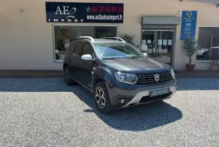 Dacia Duster gris en 3/4 avant droit, avec jantes alliage et barres de toit, stationné devant un garage AE2 Autos.