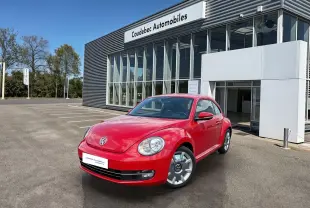 Volkswagen Coccinelle rouge vue en 3/4 avant droit, stationnée devant un bâtiment commercial moderne.