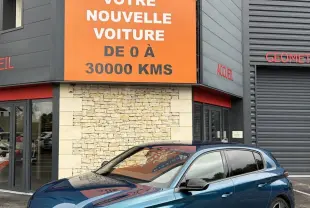Peugeot 308 bleu 3/4 avant droit, avec feux LED et jantes alliage, devant un bâtiment commercial.