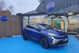 Renault Captur 2025 Bleu Iron en 3/4 avant droit, avec jantes alu noires et feux LED allumés, devant un showroom Distinxion.