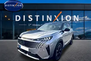 Peugeot 3008 gris artense en 3/4 avant droit, avec calandre distinctive et phares LED allumés, devant un showroom Distinxion.