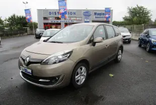 Renault Grand Scenic III beige vu en 3/4 avant droit sur parking humide devant concession automobile.