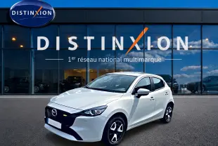 Vue 3/4 avant droite d'une Mazda2 blanche 2024 avec jantes noires et calandre noire devant un showroom Distinxion.