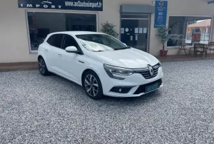 Renault Mégane IV blanc vue 3/4 avant droit, avec jantes alliage et calandre chromée, stationnée sur gravier devant un commerce.