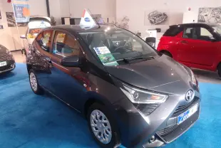 Toyota Aygo gris atlas métal vue 3/4 avant droit dans un showroom avec sol bleu et affiches promotionnelles.