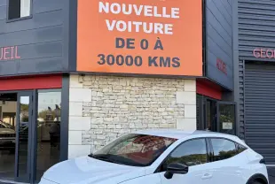 Renault Clio blanche vue en 3/4 avant droit, avec calandre noire et jantes au design moderne, devant un bâtiment commercial.