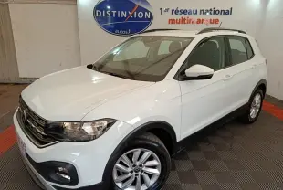 Volkswagen T-Cross blanc vue 3/4 avant droit, avec jantes alliage et barres de toit noires visibles en intérieur showroom.