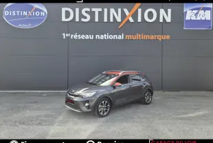 Vue 3/4 avant droite d'un Kia Stonic gris avec toit et rétroviseurs orange devant un mur gris.