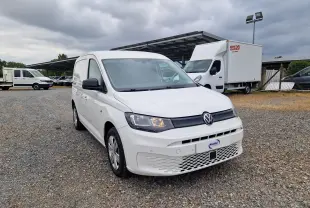 Volkswagen Caddy Cargo blanc vu en 3/4 avant droit sur un sol gravillonné avec plusieurs utilitaires en arrière-plan.