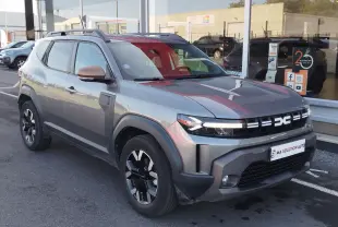Vue 3/4 avant d'un Dacia Duster 2024 gris avec rétroviseurs couleur beige, garé devant un showroom par temps de pluie.