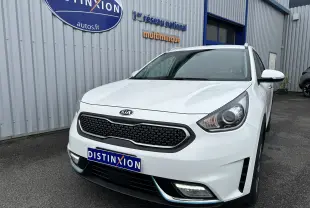 Vue 3/4 avant d'un Kia Niro hybride rechargeable blanc céleste, avec calandre noire et feux LED visibles, stationné devant un bâtiment.