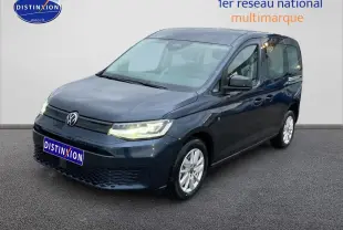 Volkswagen Caddy bleu métallisé vu de 3/4 avant droit, phares allumés sur fond neutre intérieur.