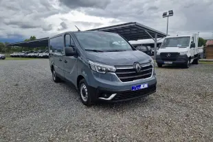 Renault Trafic Fourgon gris urbain vu en 3/4 avant droit sur un parking gravillonné avec plusieurs véhicules en arrière-plan.