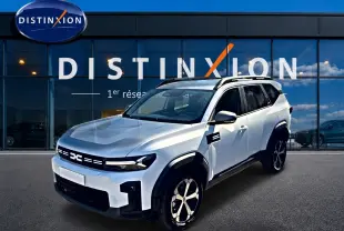 Dacia Bigster blanc en 3/4 avant droit, avec jantes alu et barres de toit visibles devant un showroom Distinxion.