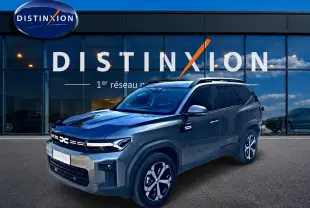Dacia Bigster gris schiste en 3/4 avant droit, avec jantes alu et barres de toit devant un showroom Distinxion.