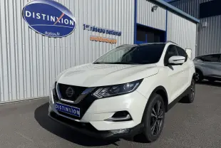 Nissan Qashqai blanc en 3/4 avant droit, avec jantes noires et calandre chromée, stationné devant un bâtiment industriel.
