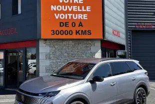 Peugeot 5008 gris clair en 3/4 avant droit, avec calandre distinctive et jantes noires, devant un bâtiment commercial.