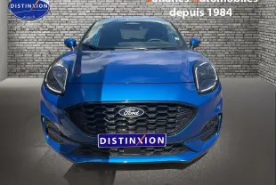 Vue frontale d'une Ford Puma bleu métallisé avec calandre noire et phares LED, sur fond de mur en briques grises.