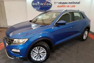 Volkswagen T-Roc bleu en 3/4 avant droit, avec jantes argentées et barres de toit blanches visibles en intérieur showroom.