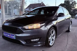 Vue 3/4 avant droite d’une Ford Focus 1.5 TDCI 120 ST Line grise avec jantes alliage 18 pouces et phares allumés.