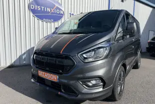 Vue 3/4 avant droit d’un Ford Transit Custom gris avec bandes noires et orange, calandre marquée Ford, devant un bâtiment.
