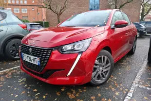 Peugeot 208 rouge Elixir vue en 3/4 avant droit, avec calandre noire et feux LED distinctifs sous la pluie.