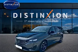 Peugeot 308 gris 3/4 avant droit, jantes alu noires, devant vitrine Distinxion avec reflet ciel bleu.