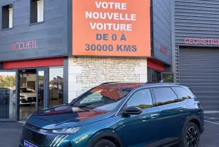 Peugeot 5008 hybride vert en 3/4 avant droit, avec jantes noires et calandre distinctive, devant un bâtiment commercial.