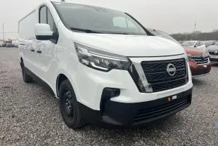 Nissan Primastar blanc en vue 3/4 avant droit sur parking extérieur avec abri métallique en arrière-plan.